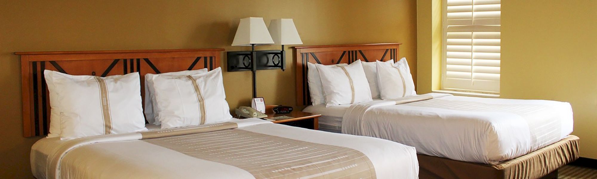 Two neatly made hotel beds with white linens, a shared nightstand, lamps, and a window with shutters in a warm-toned room.