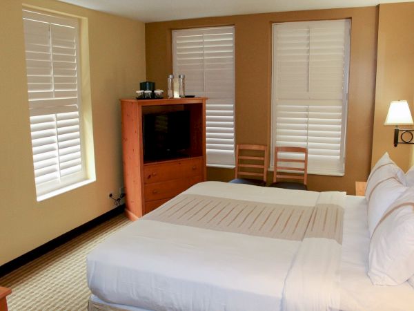 A tidy hotel room with two queen beds, a wooden dresser with a TV, and large windows with white blinds&mdash;bright and relaxing.