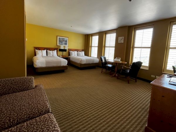 A bright hotel room with two queen beds, yellow walls, large windows, a loveseat, a desk and chairs, and a dresser facing a TV, all lit by natural light.