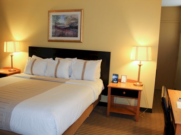 A tidy hotel room with a large bed, white linens and pillows, two bedside tables with lamps, a framed wall art, and a desk area to the right.
