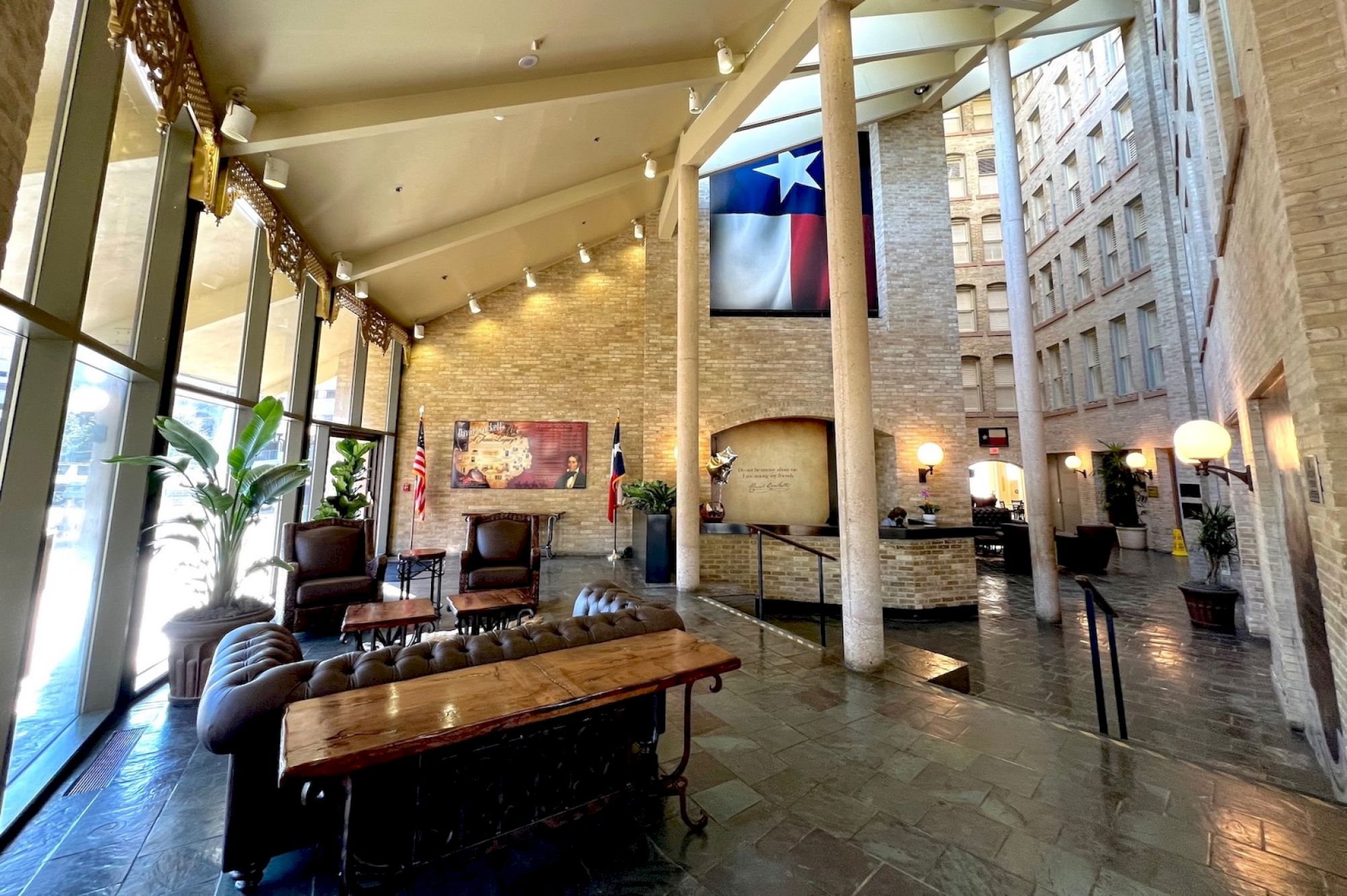 A spacious hotel lobby withstone floors, modern seating, tall windows, warm lighting, and a Texas flag hanging high.