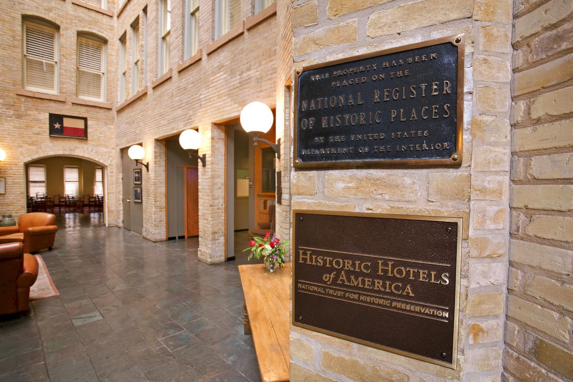 A historic hotel lobby with brick walls, hanging lamps, and plaques reading &ldquo;National Register of Historic Places&rdquo; and &ldquo;Historic Hotels of America.&rdquo;