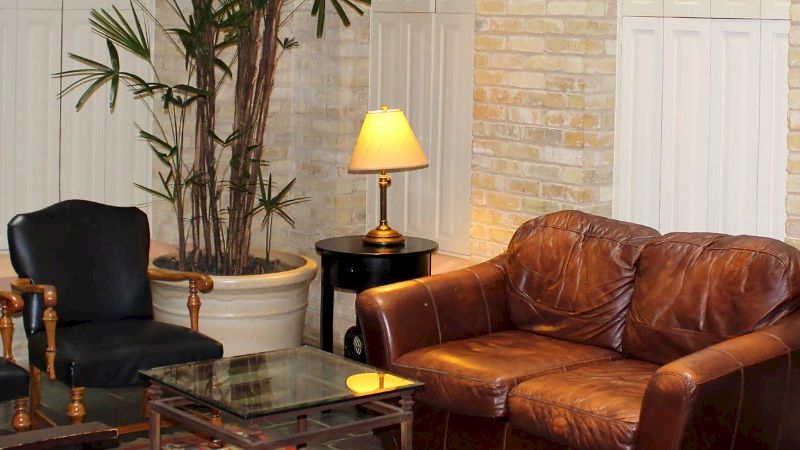 A cozy lobby or waiting area with a brown leather sofa, two black chairs, glass coffee table, lamp, plant, and framed art on a light brick wall.