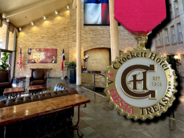 Lobby of the Crockett Hotel with a grand desk, warm lighting, a Texas flag, a large crest logo, stone walls, and upscale seating.