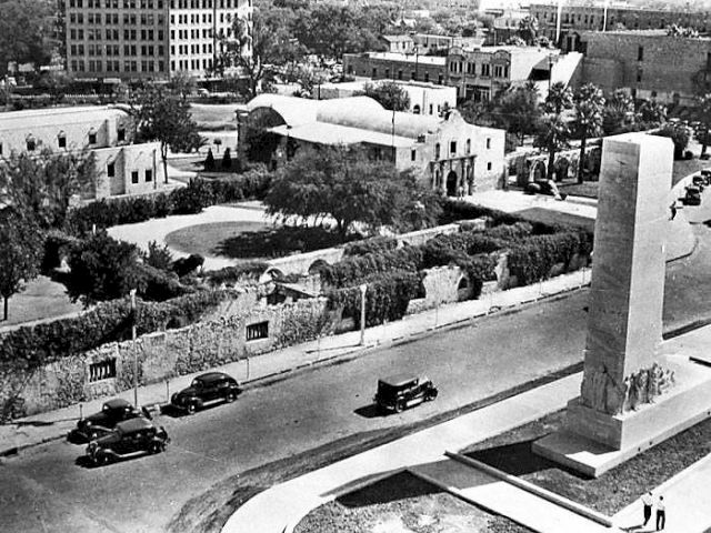 A black-and-white cityscape with a curving road, cars, low buildings, and a tall monument in the foreground, all viewed from above.