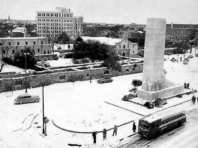Black-and-white historical street scene with a tall monument, a few cars and a bus, low buildings, and an obelisk-like structure at a snowy intersection. Top it at 140 characters, always ending the sentence.