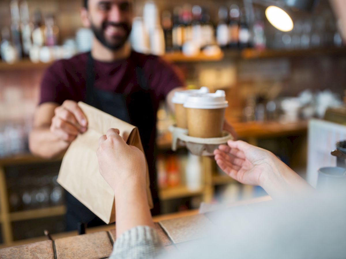 A barista hands a to-go coffee cup to a customer at a counter, with a paper bag in the foreground and shelves of bottles in the background.