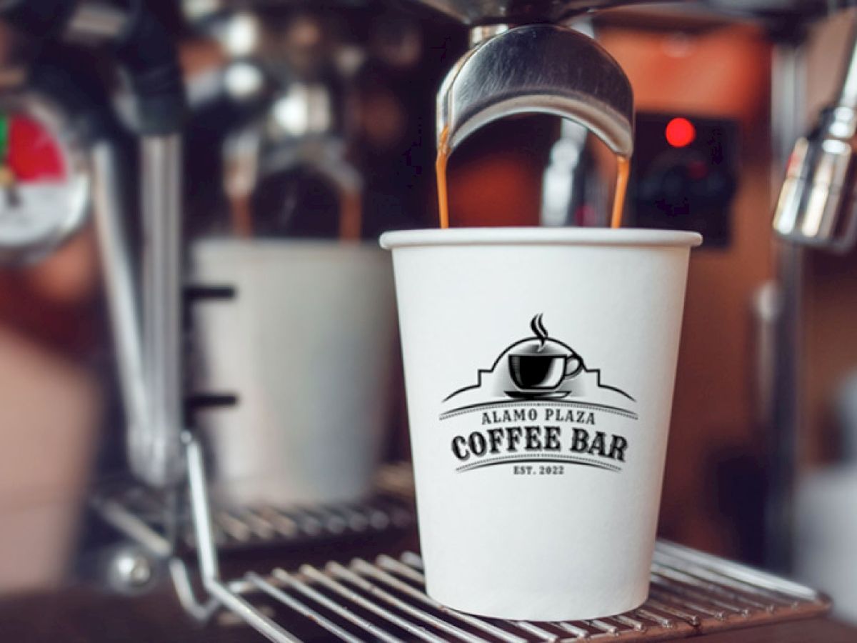 A white coffee cup with &ldquo;Alamo Plaza Coffee Bar&rdquo; logo sits under a coffee machine nozzle, ready to brew, against metal equipment.