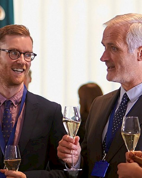 A group of professionals in business attire mingle at a social event, holding drinks and chatting warmly in a bright, modern setting.