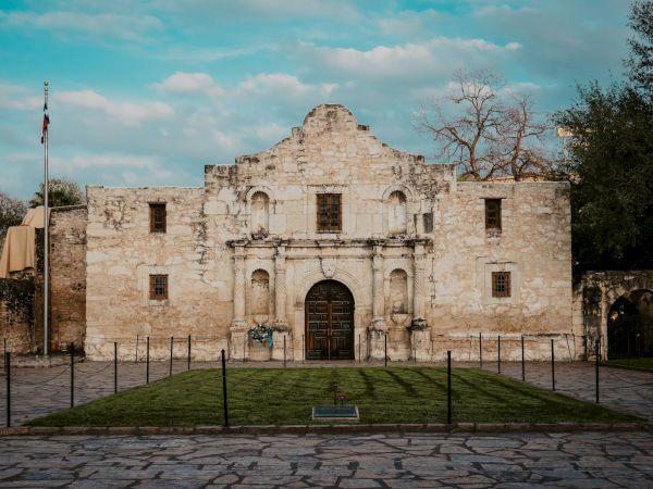 A historic stone facade with a central arched doorway, flanked by small windows, statues near the entrance, and a manicured lawn enclosed by a low fence, under a blue sky.