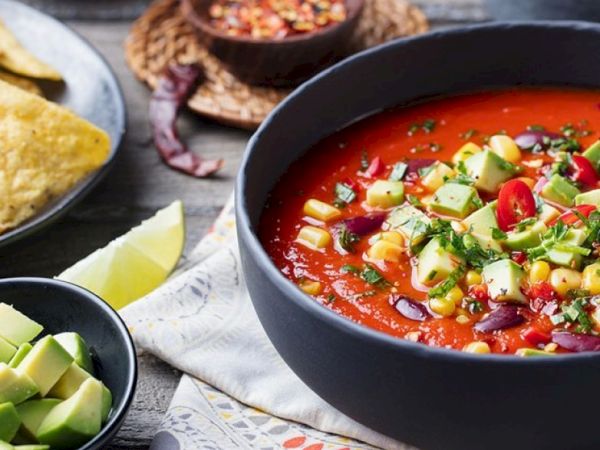 A vibrant bowl of tomato-based soup with corn, avocado, cucumber, chili peppers, and cilantro garnish, surrounded by tortilla chips and lime wedges.