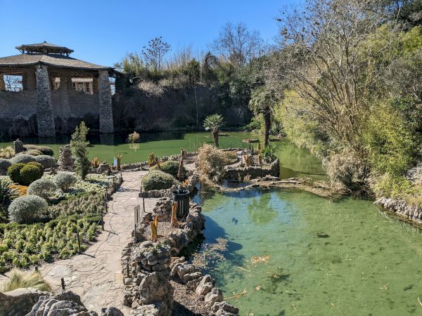 A tranquil garden with a stone path over a small pond, manicured shrubs, and a rustic building in the background, under a clear blue sky.