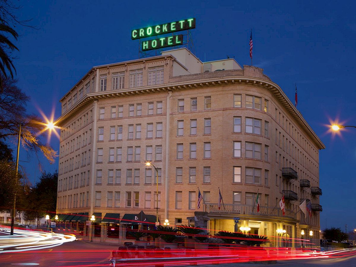 A historic brick hotel at dusk with &ldquo;Crockett Hotel&rdquo; sign on top, street lights, and light trails from passing cars.