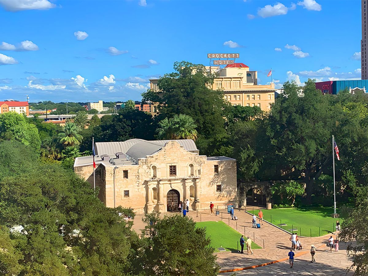 A historic stone gate and lush green park with trees, people strolling, and a bright blue sky with fluffy clouds.