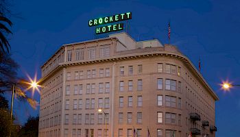 Crockett Hotel building at dusk with lit windows, sign atop, palm trees nearby, and light trails from passing cars along the street.