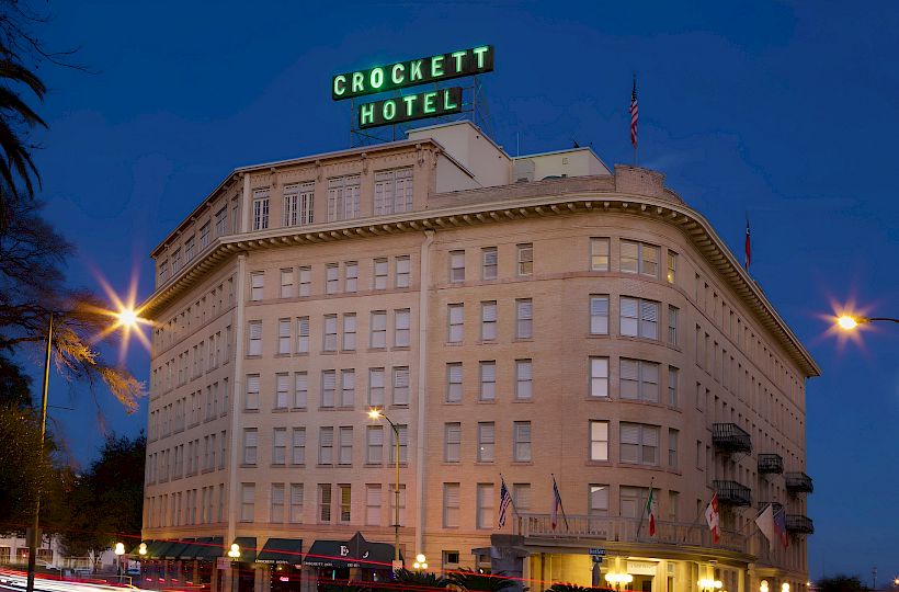 Crockett Hotel building at dusk with lit windows, sign atop, palm trees nearby, and light trails from passing cars along the street.