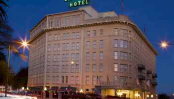 A large historic hotel at dusk with a neon &ldquo;Crockett Hotel&rdquo; sign on top, curved corner windows, and street traffic lights glowing.