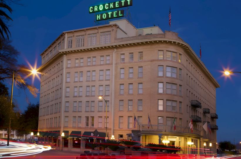 A large historic hotel at dusk with a neon &ldquo;Crockett Hotel&rdquo; sign on top, curved corner windows, and street traffic lights glowing.