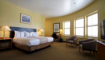 A hotel room with a large bed, two bedside lamps, a chair and desk by windows, and a dresser with a TV, all in warm tones, under soft lighting.