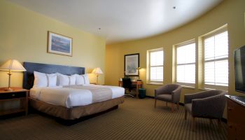 A hotel room with a large bed, two nightstands, lamps, a desk, two chairs, a dresser with a TV, and three windows letting in light.