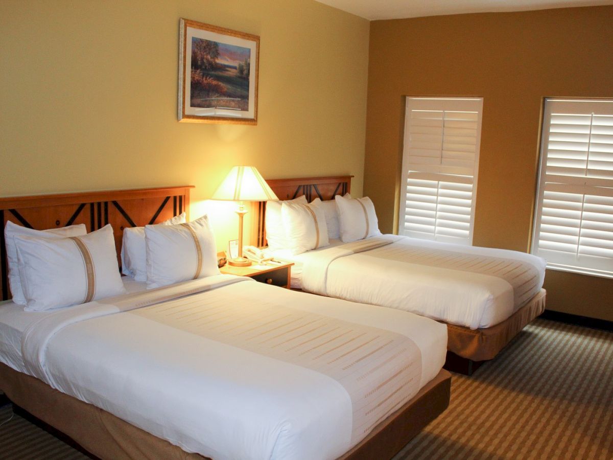 A hotel room with two double beds, nightstand with lamp, framed art, and beige walls with white blinds&mdash;cozy and tidy.