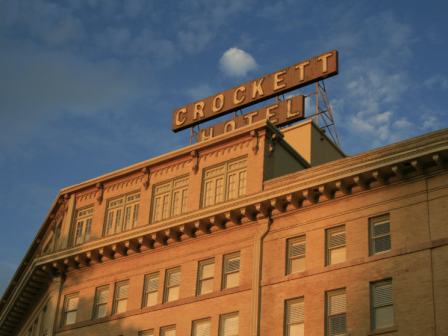 A vintage brick hotel with a bold sign reading &ldquo;Crockett Hotel&rdquo; atop its classic skyline, under a blue sky with scattered clouds and a flag.