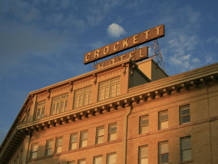 A brick hotel with a large rooftop sign reading &ldquo;CROCKETT HOTEL&rdquo; against a blue sky with a flag on a pole atop the building.