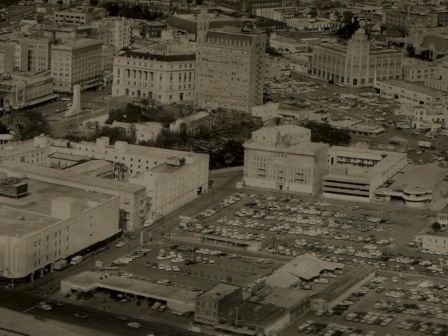 Aerial view of a mid-20th-century city with dense low-rise buildings, parking lots, and a grid-like street layout, in black and white.