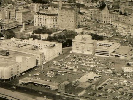 A vintage cityscape with mid‑century buildings, a large courthouse, and a parking lot full of cars, seen from above, in black and white.