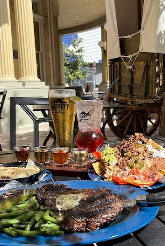 A rustic outdoor meal: two blue plates with steak, green beans, and rice; plus drinks and sauces on a wooden table near a wagon and columns.