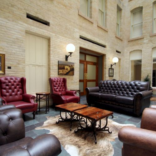 A cozy, upscale lounge with tufted leather chairs, a sofa, a wooden coffee table, warm lamps, and paintings in a brick-walled atrium.