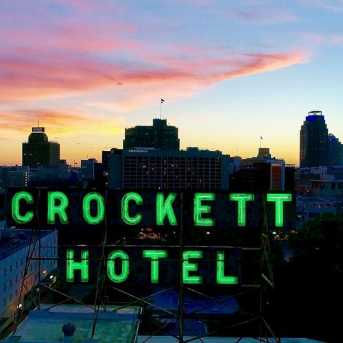 A city skyline at sunset with a neon green &ldquo;CROCKETT HOTEL&rdquo; sign perched above buildings, glowing against pink-orange sky and fading blue.