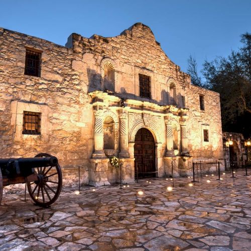 Ancient stone building with arched doorway and carved facade, old wooden cart in foreground, cobblestone ground, evening sky, trees nearby.