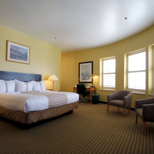 A hotel room with a large bed, two bedside lamps, a desk area, and a sitting nook by windows; warm yellow walls and striped carpet.