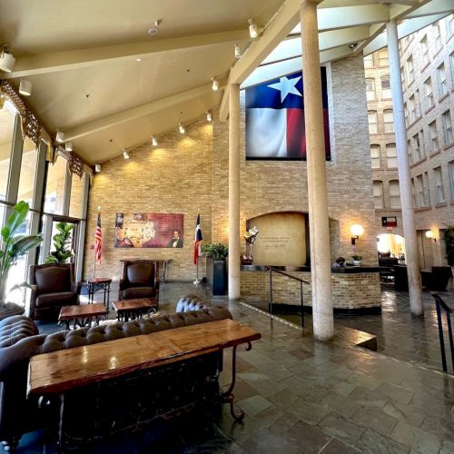 A stylish lobby with tall glass windows, a long wooden table, lounge seating, warm lighting, and a Texas flag hanging high above the stone courtyard.