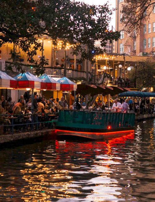 A lively riverside scene at dusk with outdoor tables along the water, a boat glowing red in the canal, and people dining under lights.