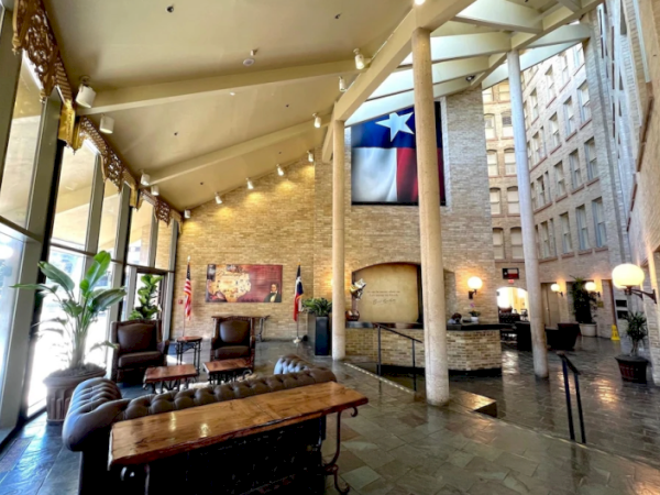 A spacious, stylish lobby or hotel atrium with tall windows, seating, plants, chandeliers, and a large flag hanging on the wall, welcoming vibe.