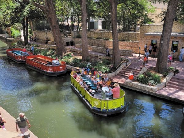 Colorful tourist boats glide along a narrow canal lined with trees and stone walls, while passengers enjoy the ride and nearby pedestrians stroll.