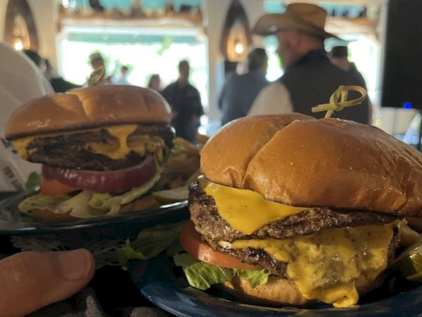Two juicy burgers with melted cheese and lettuce on sesame buns, ready to be enjoyed, in a busy restaurant setting.