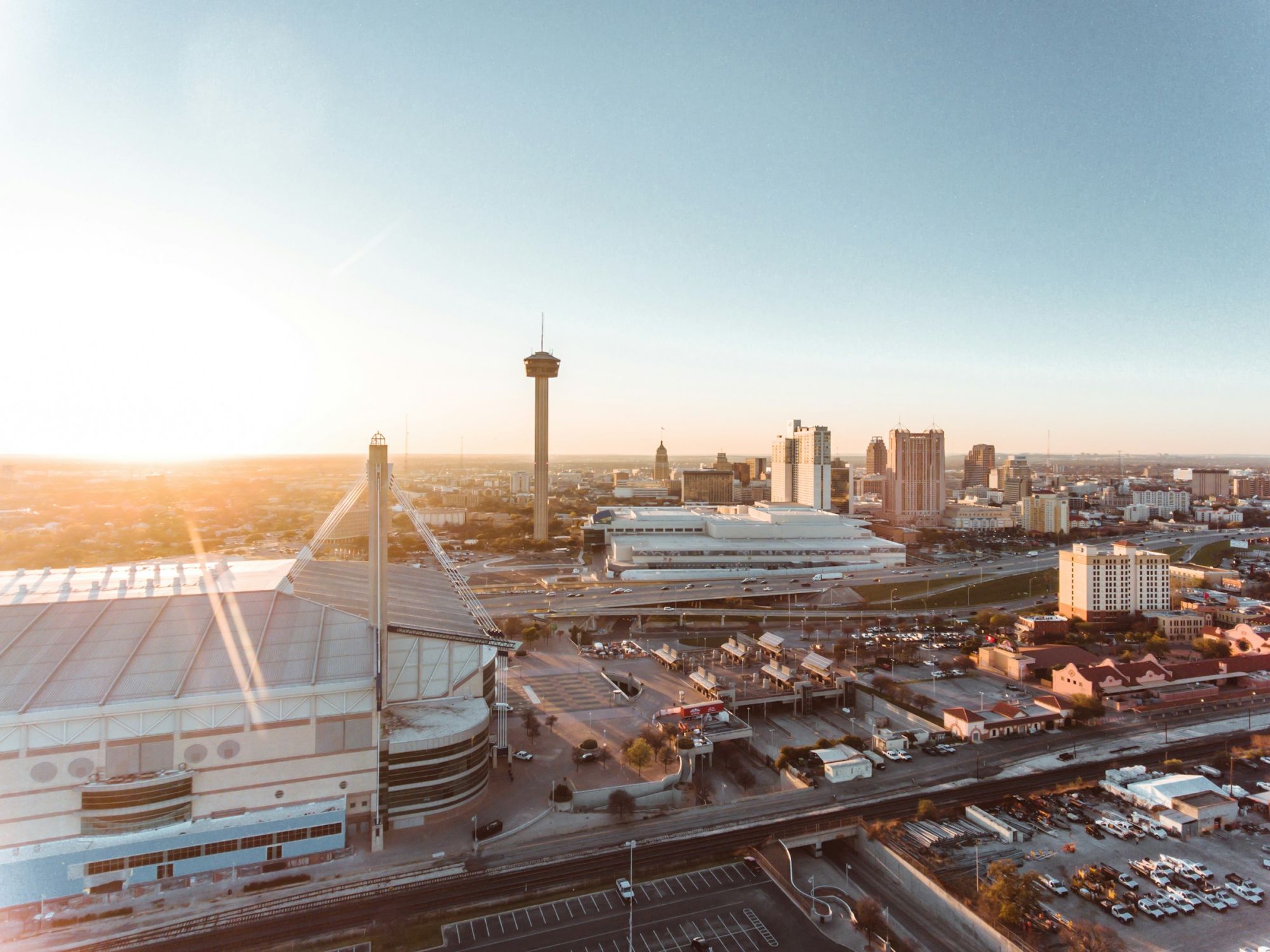 San Antonio skyline at sunset with the Alamodome, tall the tower, river, and highway, casting warm light over buildings and streets.