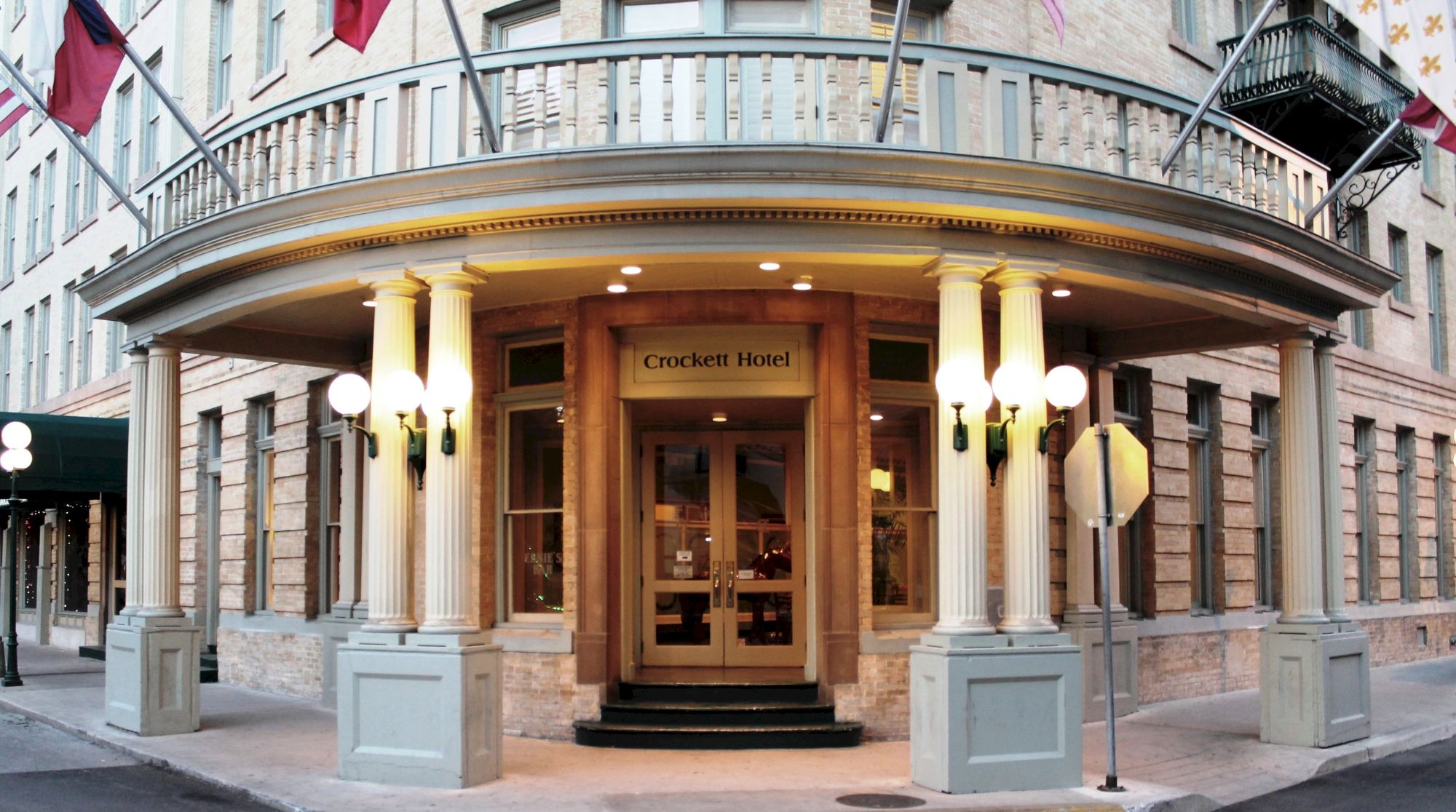 A grand hotel entrance with a curved portico, white columns, double doors, and small lights flanking the doors, city street backdrop.