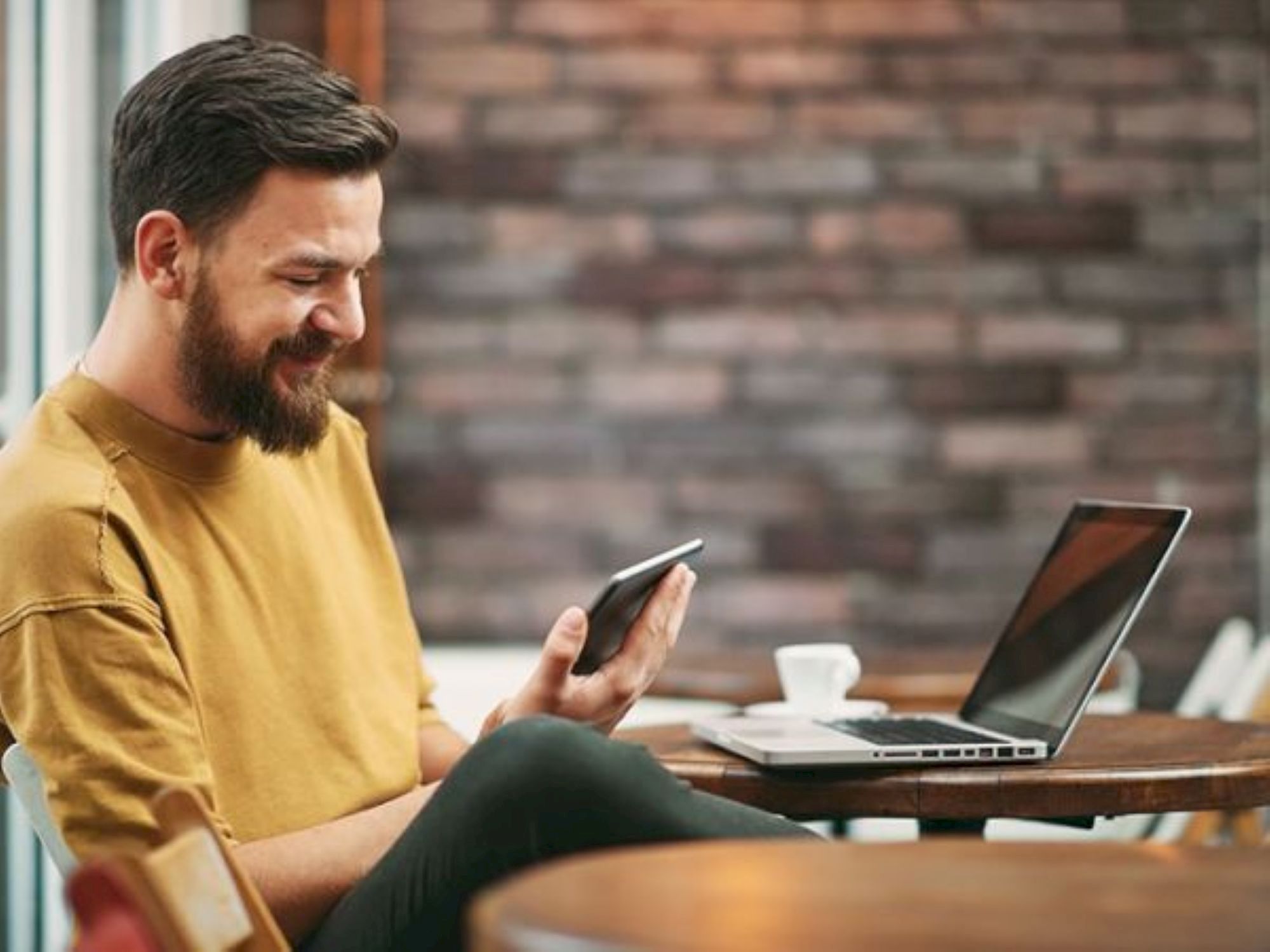 A man with a beard sits at a table with a laptop, using a smartphone, smiling, in a cozy caf&eacute; setting.