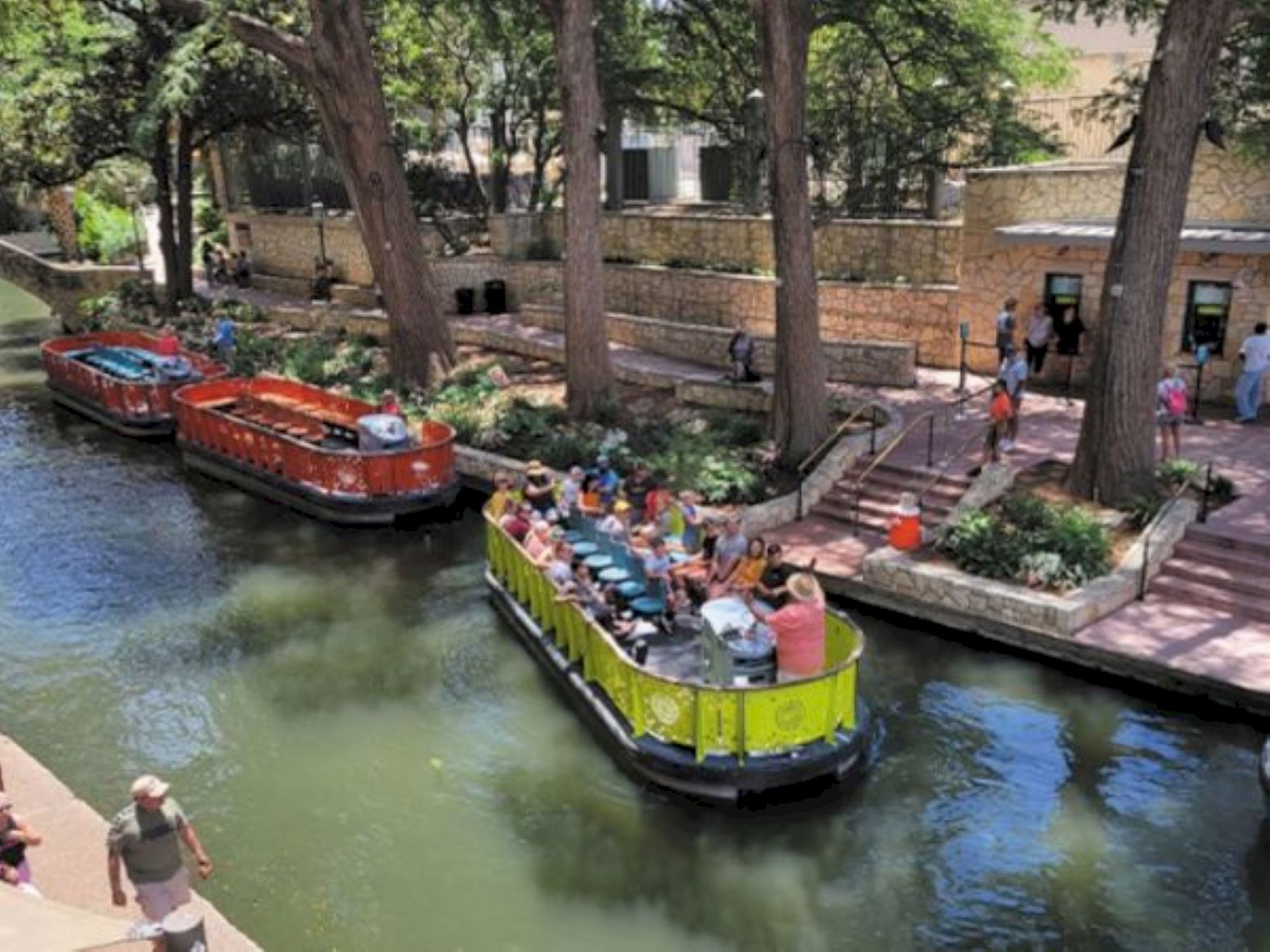 Three colorful canal boats tour a sunny urban river, people onboard enjoying the ride, while riverwalk pedestrians stroll nearby.