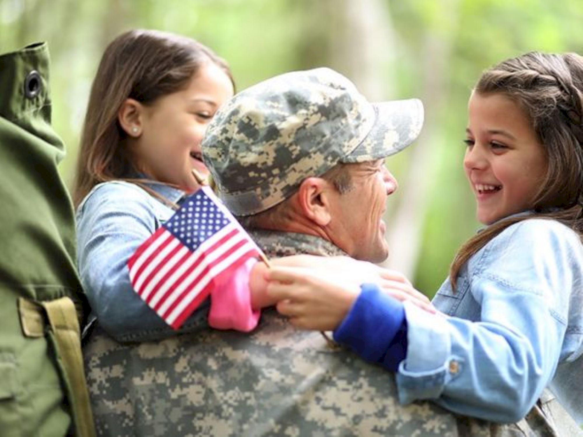 A soldier in uniform joyfully hugs two smiling children, one holding an American flag, in an outdoor setting.