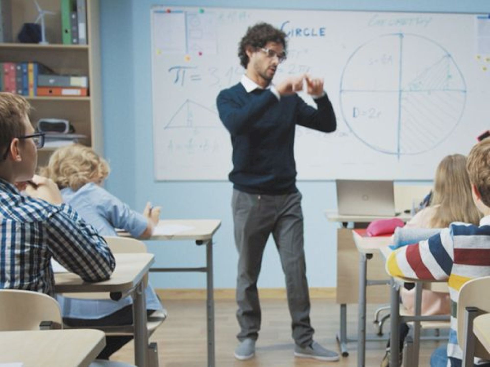 A teacher stands at the front of a classroom, gesturing as students sit at desks with a whiteboard full of math diagrams behind him.