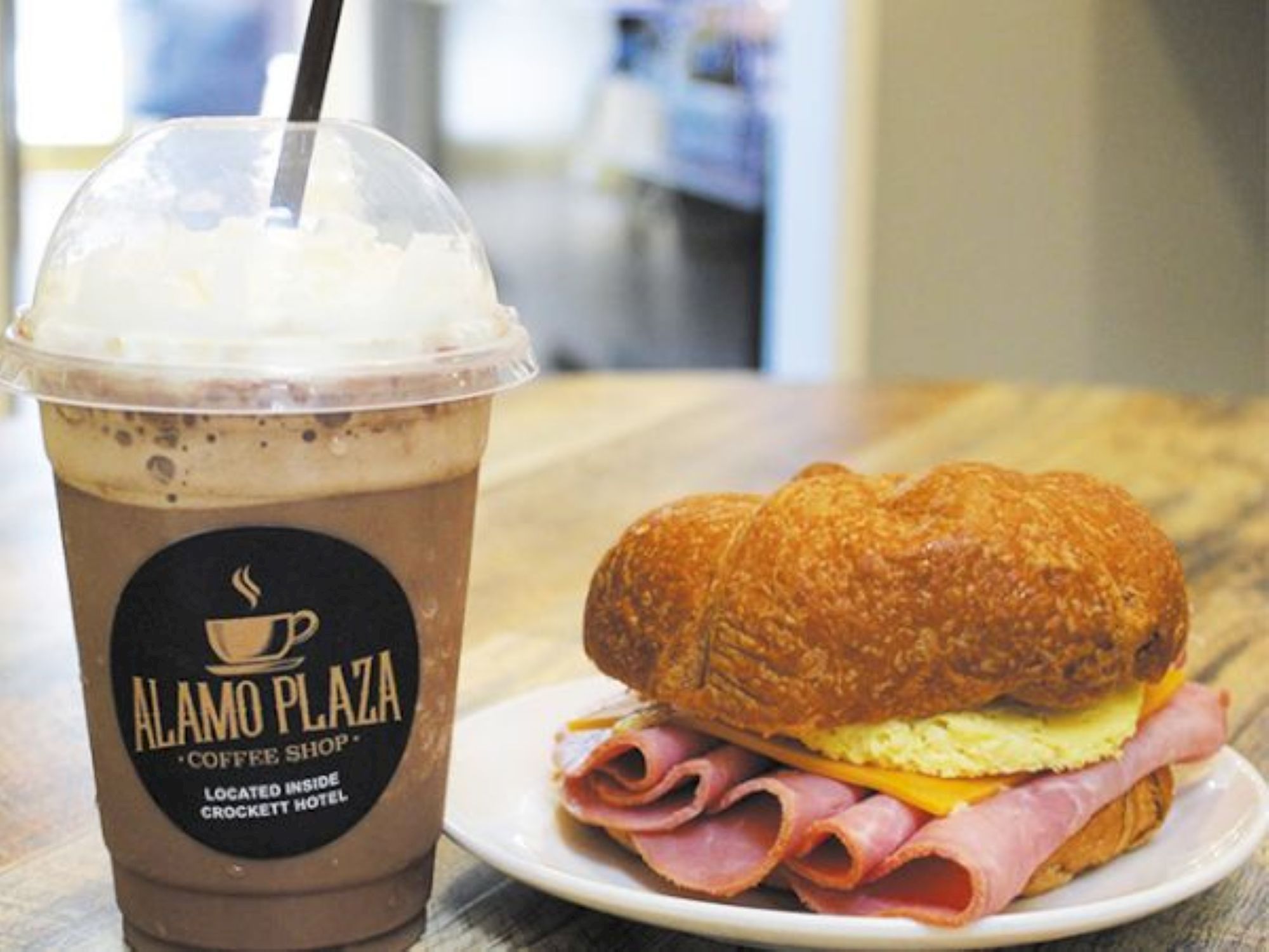 A ham and cheese croissant with sliced ham on a plate beside a cold blended coffee drink at a caf&eacute;, logo reads &ldquo;ALAMO PLAZA&rdquo; on cup.