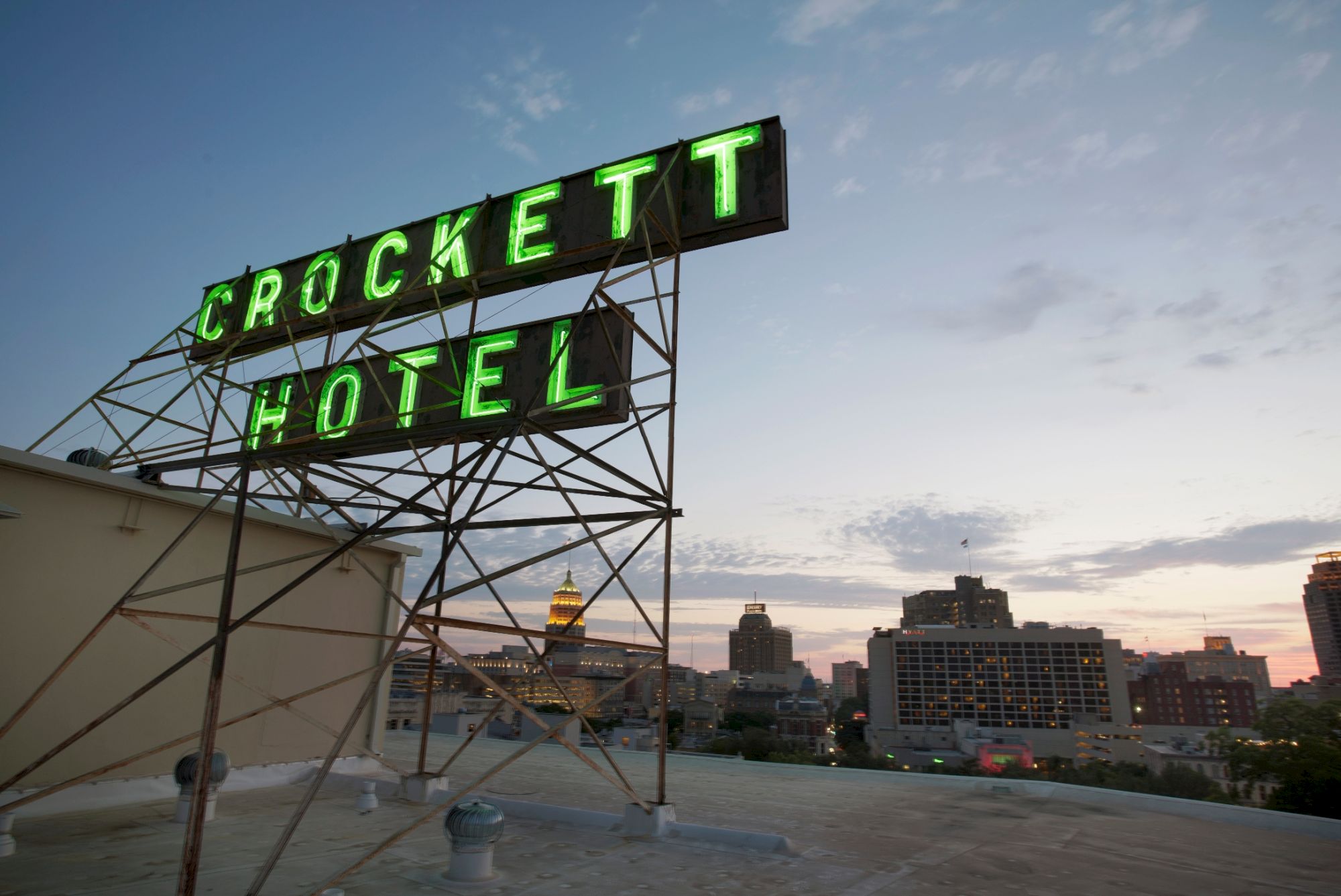 A rooftop view of the green neon &ldquo;CROCKETT HOTEL&rdquo; sign against a dawn sky over a city skyline.