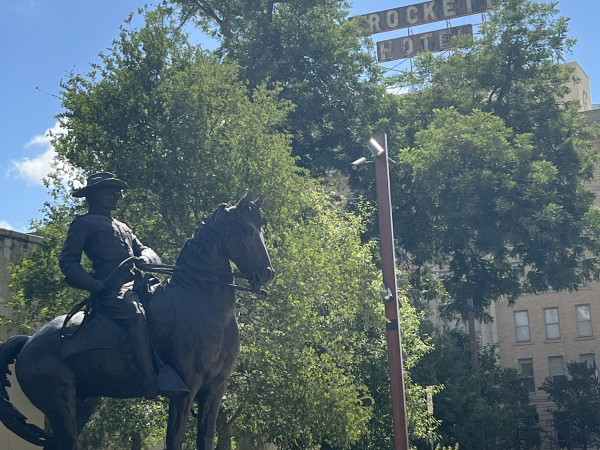 A statue of a rider on a black horse stands on a stone pedestal, with trees and a building in the background, and a sign reading &ldquo;ROCKET&rdquo; overhead.