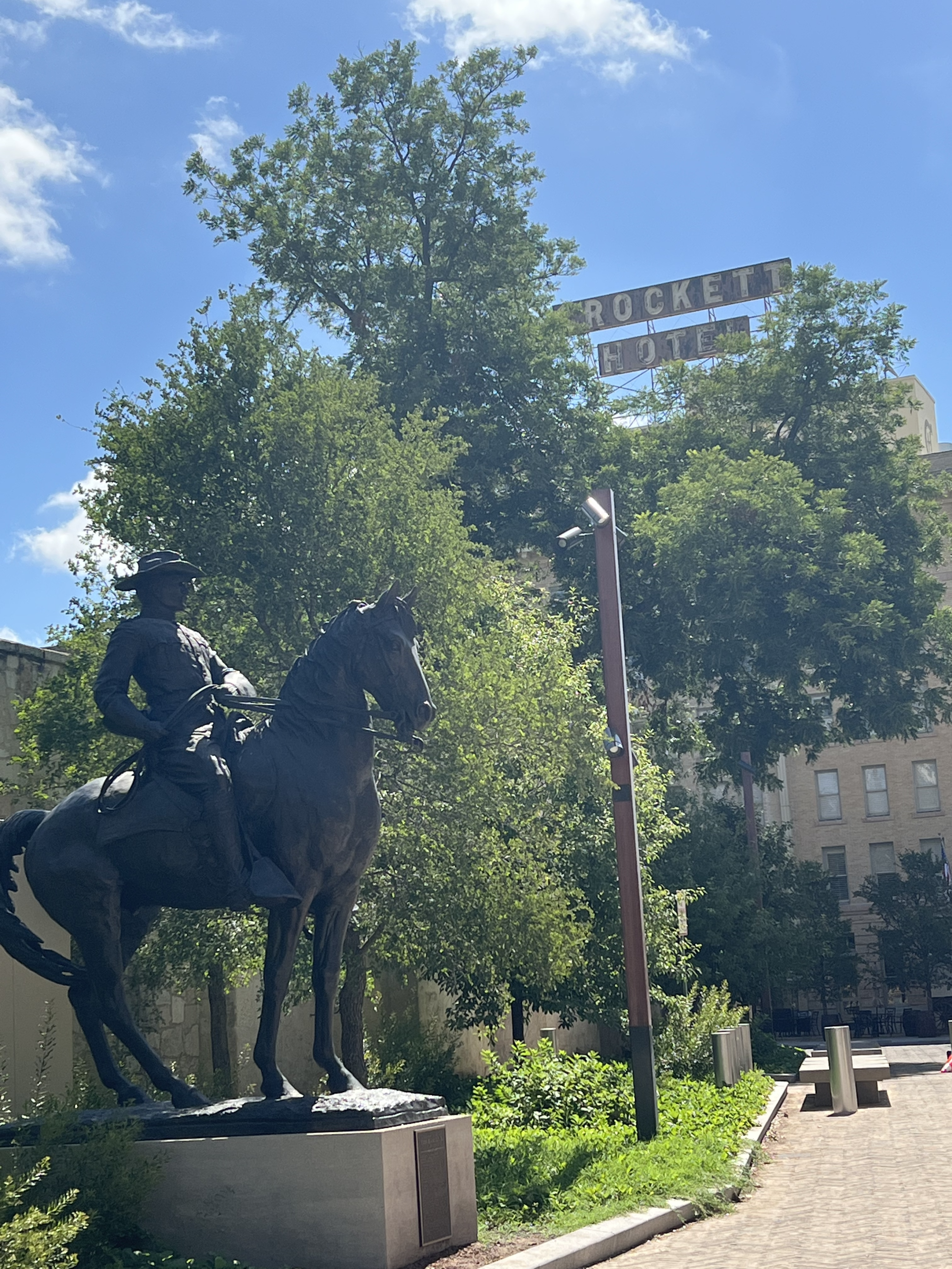 Statue of a rider on a dark horse in a sunny plaza, with green trees, a sign reading &ldquo;ROCKET&hellip;&rdquo; above, and a light brick building in the background.