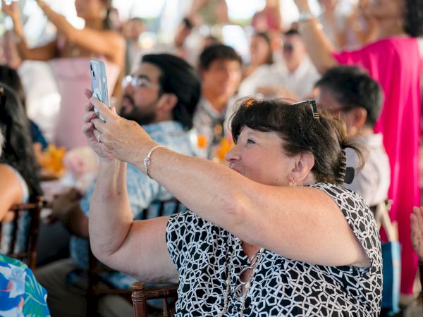 A woman in a patterned blouse takes a photo with her phone at a crowded event, others cheering in the background.
